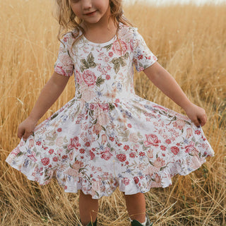 Posh Peanut Ruffled Twirl Dress - Kristiana 
A young girl wearing a short-sleeve white dress with a pink and green floral print, holding out the sides of the skirt to show its volume, standing in a field of dry golden grass.
