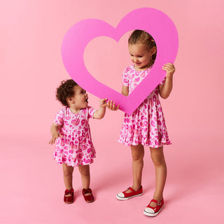 Posh Peanut Ruffled Twirl Dress - Leopard Love
Posh Peanut Ruffled Twirl Bodysuit Dress - Love Leopard
Two children in pink dresses holding a heart-shaped frame against a pink background