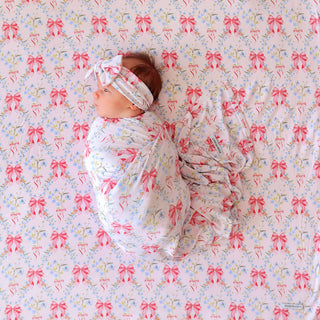 Posh Peanut Swaddle Headband Set - Harriet
A baby swaddled in a white blanket with a pink bow and blue floral print, lying on a matching printed sheet. The baby is also wearing a matching pink bow headband.