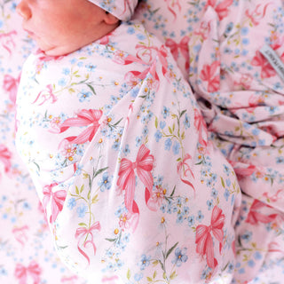 Posh Peanut Swaddle Headband Set - Harriet
A close-up of a baby swaddled in a light pink muslin swaddle blanket featuring a watercolor print of pink bows, blue forget-me-nots, and green stems. A baby's face is partially visible at the top left.