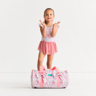 Posh Peanut Twirl Skirt Leotard - Harriet
A young girl stands on a large matching duffel bag in a studio setting. She is wearing a light pink floral sleeveless romper with a pink ruffled skirt, and the bag has a matching floral print with a large bow and pink handles.