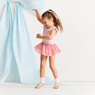 Posh Peanut Twirl Skirt Leotard - Harriet
A young girl with a pink hair tie and braid is wearing a pink and white floral sleeveless tutu dress with a matching pink skirt, white socks, and ballet slippers, posing in front of a light blue fabric backdrop.