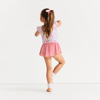 Posh Peanut Twirl Skirt Leotard - Harriet
A young girl with long brown hair in a ponytail, wearing a pink and white floral leotard with a pink skirt, posing from behind against a clean white studio background.