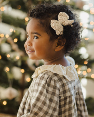 Quincy Mae Crochet Bow - Natural
Child wearing a checkered dress with a decorative headband in front of a blurred Christmas tree.