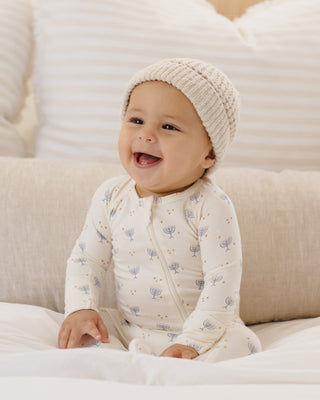 
Baby wearing a cream-colored knit hat and matching outfit sitting on a beige couch.