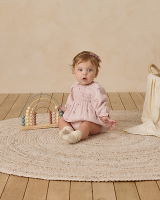 Quincy Mae Eden Romper - Bows
Baby in a pink dress sitting on a rug with a wooden toy in a neutral-colored room.