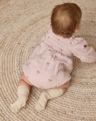 Quincy Mae Eden Romper - Bows
Baby in a pink dress with bow patterns sitting on a textured beige surface