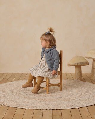 Quincy Mae Simone Dress - Blue Plaid
Child sitting on a wooden chair in a room with a beige wall and wooden floor.