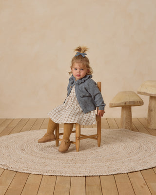 Quincy Mae Simone Dress - Blue Plaid
Young girl sitting on a wooden chair in a room with a wooden floor and beige walls.