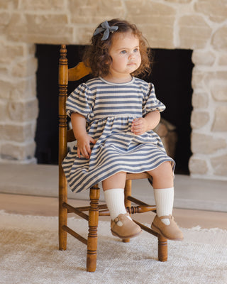 Quincy Mae Darla Dress - Ocean Stripe
Young girl in a striped dress sitting on a wooden chair in front of a stone fireplace.
