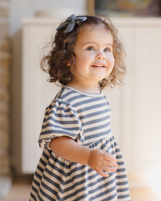 Quincy Mae Darla Dress - Ocean Stripe
Young girl in a striped dress with a bow in her hair, standing indoors.