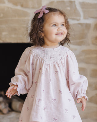Quincy Mae V Smocked Dress - Bows
Child wearing a light pink dress with bow patterns in front of a stone wall.