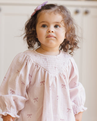 Quincy Mae V Smocked Dress - Bows
Young girl wearing a pink dress with bow patterns against a neutral background