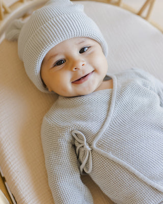 Quincy Mae Baby Bear Beanie - Ice Blue
Baby wearing a light gray knit outfit and hat, smiling on a beige surface.