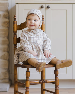 Quincy Mae Simone Dress - Blue Plaid
Child wearing a plaid dress and bonnet sitting on a wooden chair indoors.