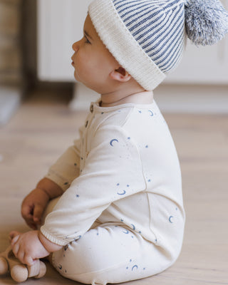 Quincy Mae Henley Bubble Jumpsuit - Moons
Baby sitting on a wooden floor wearing a white onesie with blue patterns and a striped hat.
