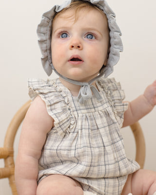 Quincy Mae Maya Romper - Blue Plaid
Baby wearing a checkered dress and gray bonnet sitting on a wooden chair.