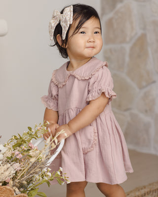 Quincy Mae Bow - Dahlia
Child wearing a pink dress with ruffles and a large bow, standing against a stone wall.