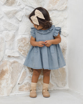 Quincy Mae Sophie Dress - Chambray
Child wearing a denim dress with ruffled sleeves against a stone wall.