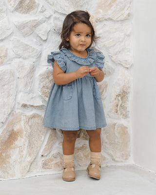 Quincy Mae Sophie Dress - Chambray
Young girl wearing a light blue dress with ruffled collar against a stone wall.
