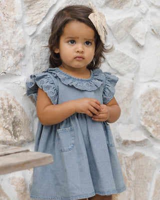 Quincy Mae Sophie Dress - Chambray
Young girl wearing a denim dress with ruffled details against a stone wall.