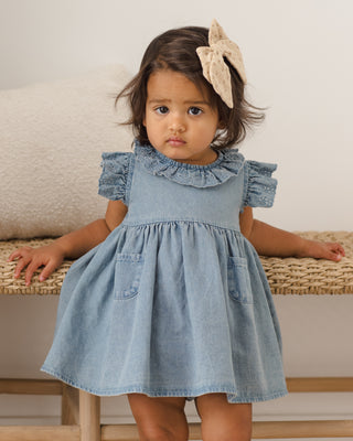 Quincy Mae Sophie Dress - Chambray
Young girl wearing a denim dress with a bow in her hair, sitting on a woven bench.
