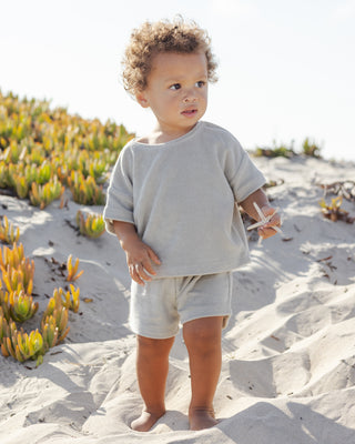 Quincy Mae Terry Top + Short Set - Sage
Child in light gray outfit standing on a sandy beach with greenery in the background