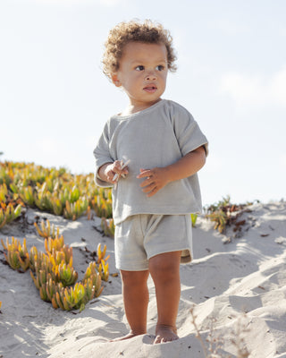 Quincy Mae Terry Top + Short Set - Sage
Child in light gray outfit standing on a sandy beach with greenery in the background