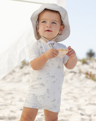 Quincy Mae Zip Rashguard One-Piece - Dinosaur
Baby in a white outfit with animal patterns and a sun hat on a beach.