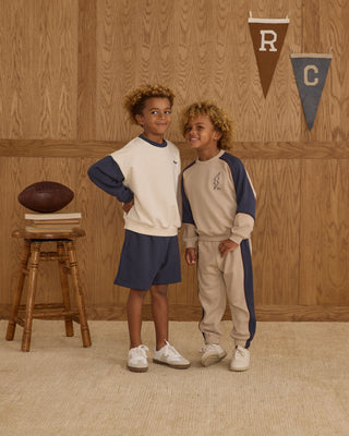 Play by Rylee & Cru Paneled Sweatpant - Stone Two children in matching outfits standing in a room with wooden walls and a stool with a football.