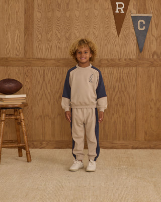 Play by Rylee & Cru Paneled Sweatshirt - Stone Child wearing a beige and navy tracksuit standing in front of a wooden wall with sports-themed decor.