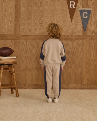 Play by Rylee & Cru Paneled Sweatshirt - Stone Child wearing a beige and navy tracksuit standing in front of a wooden wall with decorative elements.