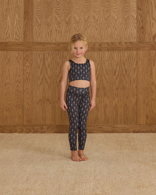 Play by Rylee & Cru Swift Sports Bra - Vintage Bloom Play by Rylee & Cru Basic Legging - Vintage Bloom Young girl wearing a navy patterned outfit standing against a wooden paneled wall.