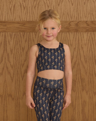 Play by Rylee & Cru Swift Sports Bra - Vintage Bloom Play by Rylee & Cru Basic Legging - Vintage Bloom Young girl wearing a navy blue floral outfit against a wooden paneled wall.