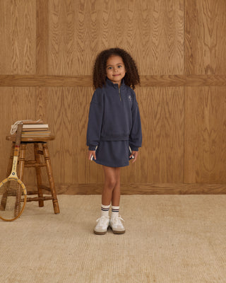 Play by Rylee & Cru Half Zip Sweatshirt - Navy Play by Rylee & Cru Sports Skirt - Navy Child wearing a navy tracksuit standing in front of a wooden paneled wall.