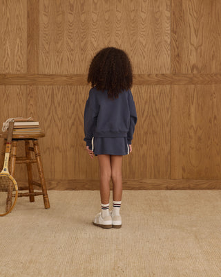 Play by Rylee & Cru Half Zip Sweatshirt - Navy Play by Rylee & Cru Sports Skirt - Navy Person wearing a navy blue outfit standing in front of a wooden paneled wall.