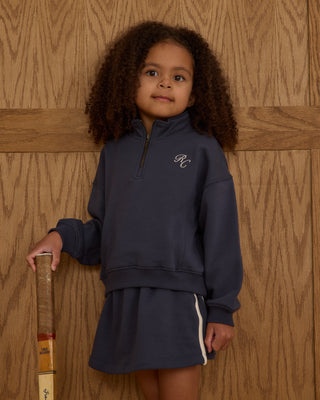 Play by Rylee & Cru Half Zip Sweatshirt - Navy Play by Rylee & Cru Sports Skirt - Navy Young girl in navy blue outfit with logo, holding a wooden object against a wooden background