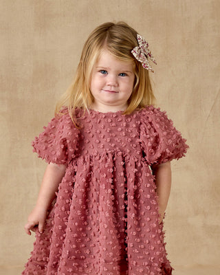 Noralee Luna Dress - Raspberry
Young girl wearing a pink dress with a textured pattern against a beige background
