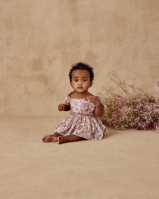 Noralee Margot Romper - Harvest Fleur
Baby in a floral dress sitting on a beige floor with flowers to the side