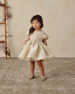 Noralee Sofia Dress - Ecru
Young girl in a beige dress standing on a textured floor with a neutral wall background