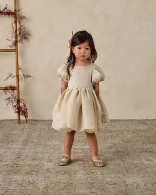 Noralee Sofia Dress - Ecru
Young girl in a beige dress standing in a room with a wooden ladder and dried plants.