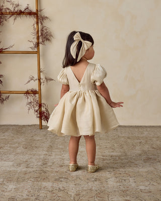 Noralee Everly Bow - Ecru
Young girl in a beige dress standing in a room with a wooden ladder and decorative plants.