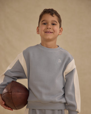 Play by Rylee & Cru Relaxed Sweatshirt - Dusty Blue
Child wearing a gray and beige sweatshirt holding a brown ball against a beige background