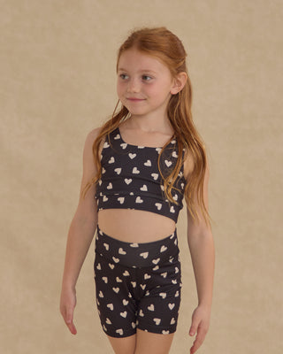 Play by Rylee & Cru Swift Sports Bra - Hearts
Young girl wearing a black tank top and shorts with heart patterns on a beige background