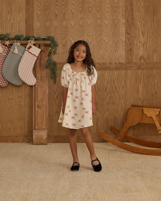 Rylee & Cru Sabrina Mini Dress - Ruby Bows
Young girl in a white dress with a pattern standing in front of a wooden wall with stockings and a rocking horse.