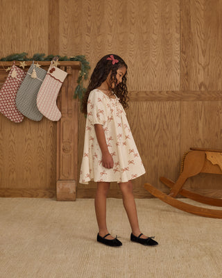 Rylee & Cru Sabrina Mini Dress - Ruby Bows
Young girl in a white dress standing in a room with wooden walls and a rocking horse.