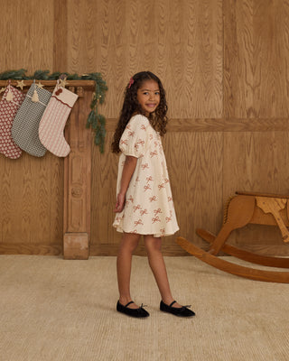 Rylee & Cru Sabrina Mini Dress - Ruby Bows
Young girl in a white dress with red patterns standing in a room with wooden walls and a rocking horse.
