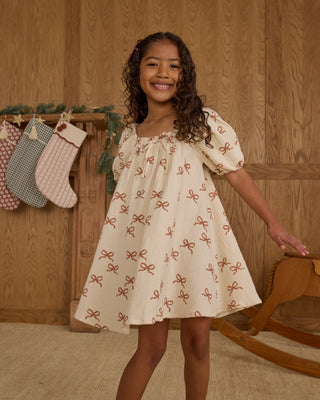 Rylee & Cru Sabrina Mini Dress - Ruby Bows
Young girl in a patterned dress standing in front of a wooden wall with stockings.