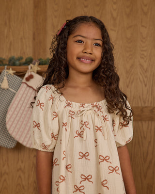 Rylee & Cru Sabrina Mini Dress - Ruby Bows
Young girl wearing a beige dress with red bow patterns against a wooden background