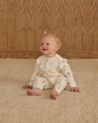 Rylee & Cru Henley Jumpsuit - Nutcracker
Baby sitting on a textured rug wearing a floral outfit with a wooden wall background